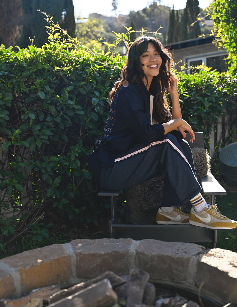Woman sitting outdoors by a stone fire pit with greenery in the background