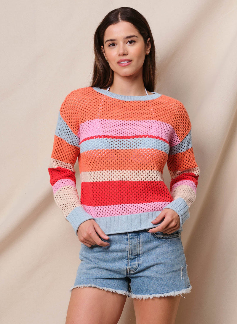 A female model wearing a striped pullover sweater with mesh detailing, in colors orange, red, pink, and off-white, paired with denim shorts.