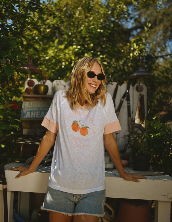 A woman smiling and wearing a white t-shirt with a graphic, blue denim shorts, sunglasses, and standing in front of a garden backdrop.
